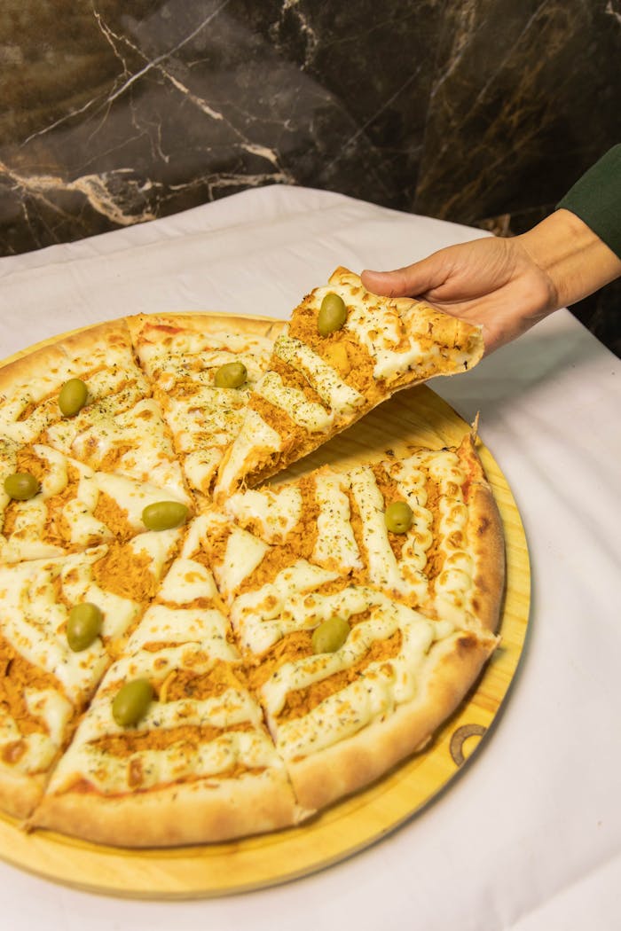 why-choose-us Close-up of a hand serving a slice of cheesy pizza topped with green olives, on a wooden board.